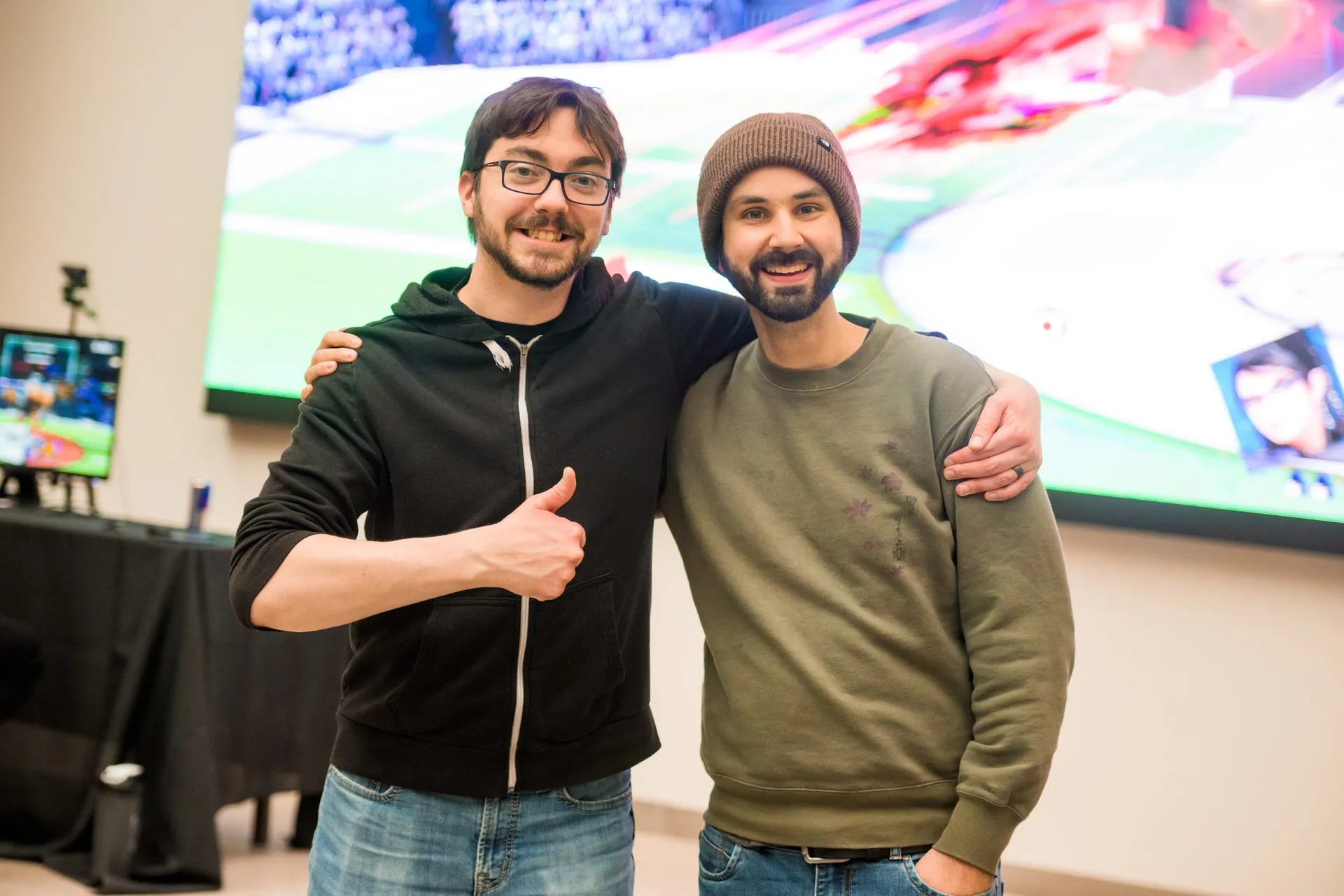 Two attendees pose with a thumbs up in front of the main screen at Canakup 2023